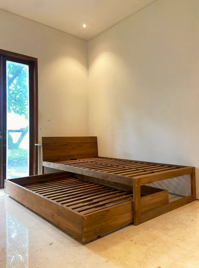 Sebuah ranjang sorong atau trundle bed modern yang terbuat dari kayu jati reclaimed, dengan ranjang bawah yang ditarik keluar sebagian, berada di sudut kamar tidur minimalis dengan lantai marmer.