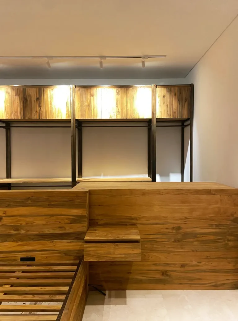 Tampilan detail headboard multifungsi dan nakas gantung dari kayu jati daur ulang, menonjolkan tekstur alami kayu dengan latar belakang lemari pakaian berbingkai industrial yang disorot oleh lampu track di atasnya.