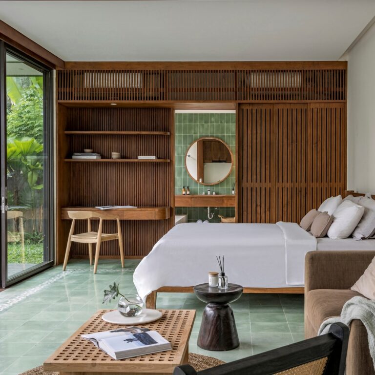 Kamar tidur berkonsep terbuka dengan furnitur kayu kustom yang menyatu, termasuk partisi kisi-kisi, meja kerja, dan ranjang, dengan area kamar mandi semi-terbuka berubin hijau dan pintu kaca besar menuju taman