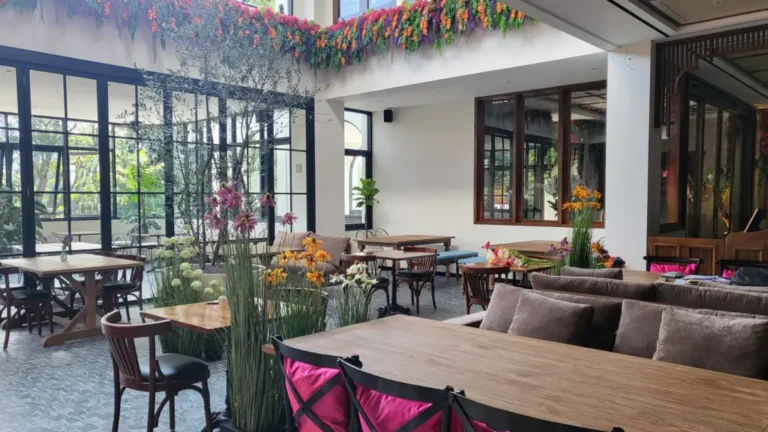 Area makan semi-outdoor di Restoran Madame Djeli dengan konsep taman, menampilkan custom furniture jati dari Permata Furni termasuk sofa panjang berwarna coklat dan meja makan kayu solid, dikelilingi dekorasi tanaman dan bunga.