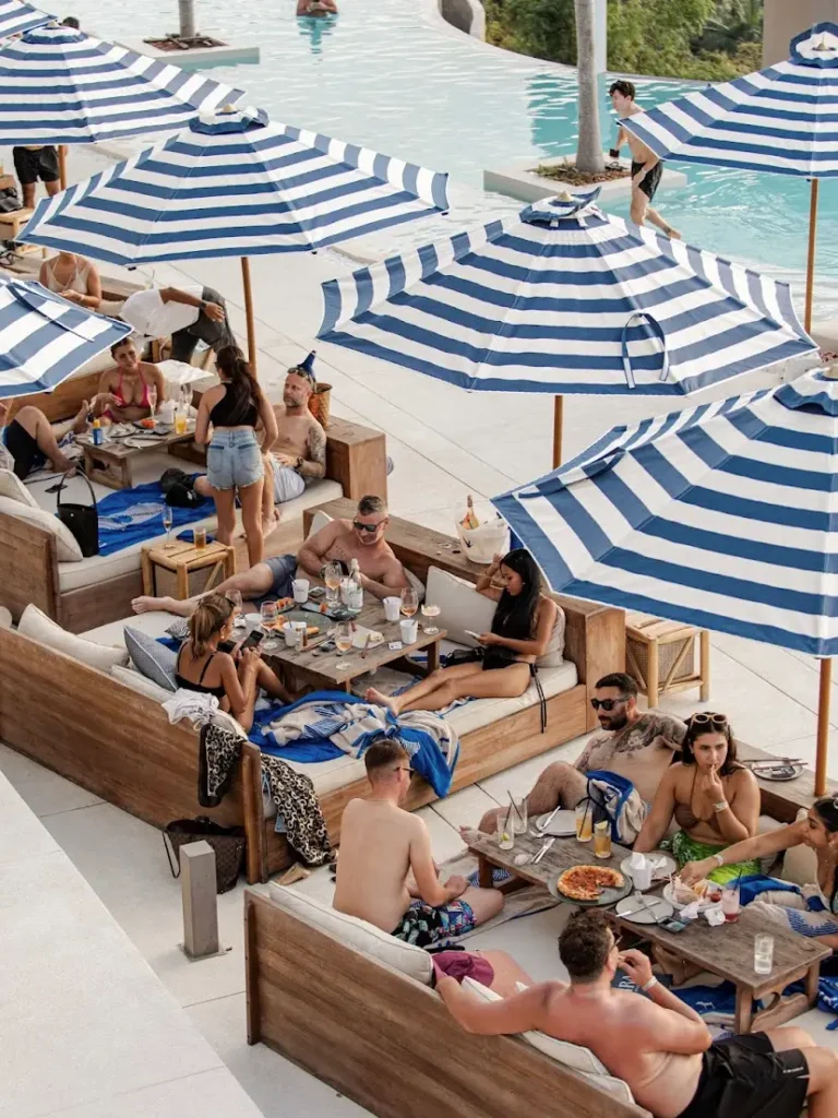 Tamu menikmati suasana Fira Beach Club di Phuket, bersantai di atas set sofa outdoor dan daybed nyaman dari kayu jati reclaimed produksi Permata Furni.